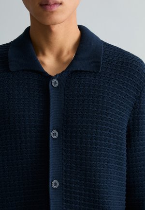 Mann trägt eine dunkelblaue, strukturierte Strickjacke mit Kragen und drei sichtbaren Knöpfen, steht vor einem schlichten hellen Hintergrund.