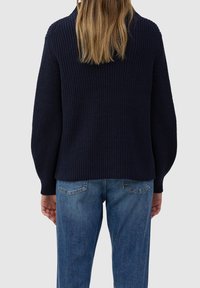 Marineblauwe gebreide trui met geribbelde textuur en verlengde schoudernaden, gecombineerd met lichtblauwe jeans, gezien van achteren.