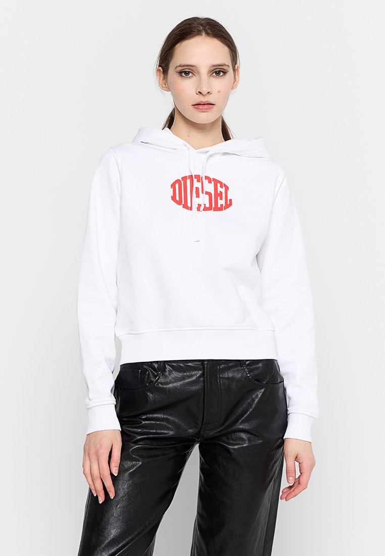 Femme portant un sweat à capuche blanc avec le logo rouge "DIESEL" et un pantalon en cuir noir, debout devant un fond blanc uni.