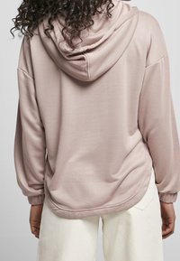 Mikina s kapucí v měkké růžové tkanině s pružnými manžetami a lemem, s uvolněným střihem a hladkou texturou. Pod mikinou jsou viditelné bílé kalhoty.
