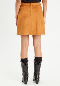 Jupe A-line en daim beige avec une fermeture éclair au dos, associée à des bottes de style western noires et des chaussettes noires côtelées.