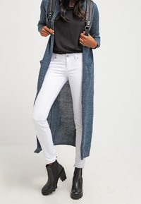Cardigan lungo blu in maglia strutturata, indossato sopra una maglietta nera, abbinato a jeans skinny bianchi e stivali neri alla caviglia.