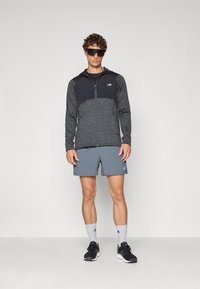 Veste athlétique légère de couleur gris foncé avec une fermeture éclair à l'avant, assortie à un short gris. Des chaussures de course noires et des chaussettes de sport complètent le look.