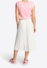 Donna in piedi di spalle, che indossa un top smanicato rosa, una gonna midi plissettata bianca e sandali piatti rosa con fiocchi.