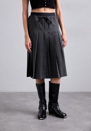 Femme portant une jupe plissée grise longueur genoux avec une taille à cordon et des bottes en cuir noires montant jusqu'aux genoux sur un sol gris.