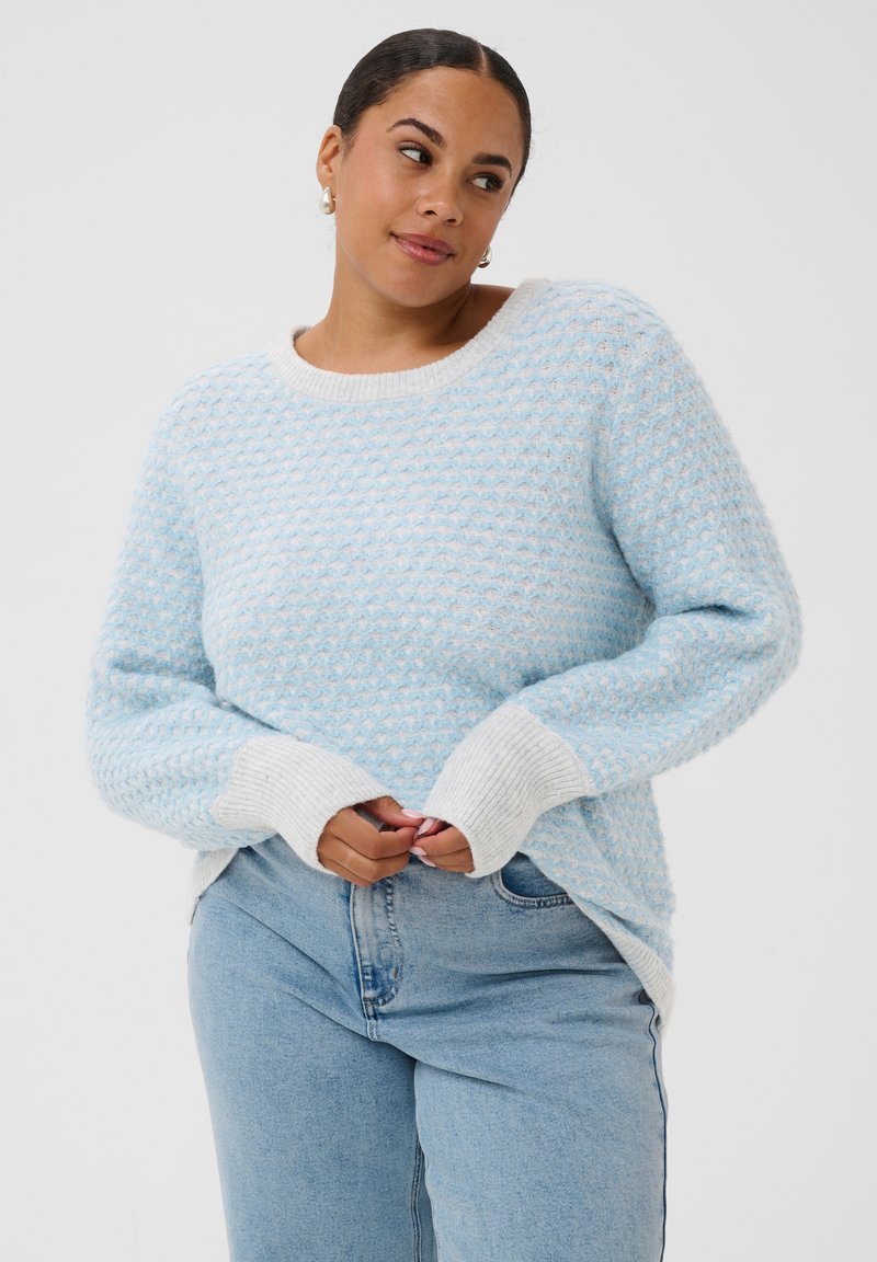 Femme portant un pull texturé bleu clair et blanc avec des manches retroussées et un jean bleu clair, debout les mains jointes et regardant de côté.