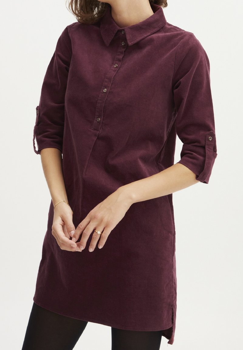 Femme portant une robe-chemise bordeaux à manches longues boutonnée avec les manches retroussées, des leggings noirs, et un bracelet et une bague en or.