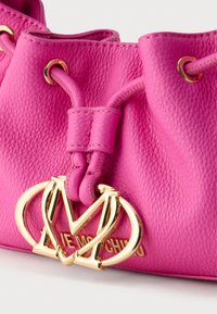 Ροζ δερμάτινη τσάντα τύπου κουβά με υφή στην επιφάνεια, διακοσμημένη με εξαρτήματα σε χρυσό τόνο και ένα έντονο λογότυπο στο μπροστινό μέρος.