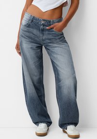 Kvinna klädd i högmidjade urtvättade vida jeans med en hand i fickan, ihop med vita sneakers och en kortärmad vit topp.