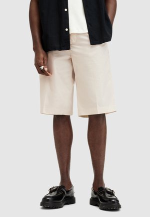 Shorts beiges en tissu léger, arrivant au genou avec une texture lisse, associés à des chaussures noires avec quincaillerie décorative.