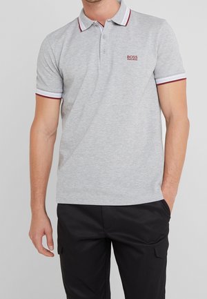 Mężczyzna w jasnoszarej koszulce polo Hugo Boss z bordowo-białym prążkowanym kołnierzem i rękawami, w połączeniu z czarnymi spodniami.