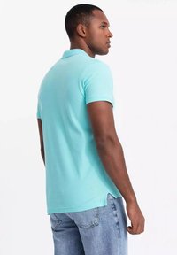 Polo triko v nebesky modré barvě vyrobené z texturované bavlny, se límcem, krátkými rukávy a postranním rozparkem na spodním lemu, v kombinaci s světlými modrými džíny.