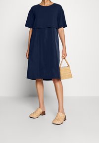 Dunkelblaues Baumwollkleid mit einem geschichteten Oberteil, kurzen Ärmeln und einer lockeren A-Linie. Eine gewebte beiges Tasche und geflochtene braune Clogs runden das Outfit ab.