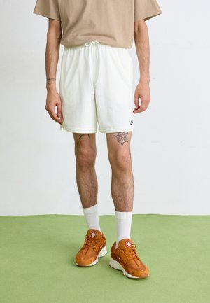 Șorturi albe cu un brâu elastic și sfoară, asortate cu un tricou bej și adidași portocalii cu tălpi albe și accente de sosete.