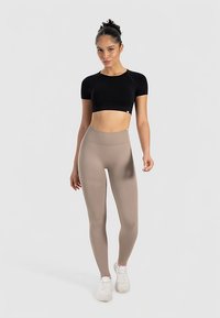 Schwarzes, kurzes Oberteil mit kurzen Ärmeln, kombiniert mit hellbeigen, hoch taillierten Leggings. Model trägt weiße Sneaker und steht vor einem neutralen Hintergrund.