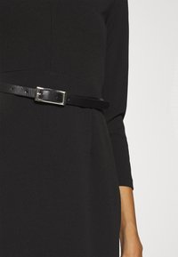 Vestido negro con mangas tres cuartos, que presenta un corte ajustado y un cinturón de cuero negro brillante con una hebilla plateada en la cintura.