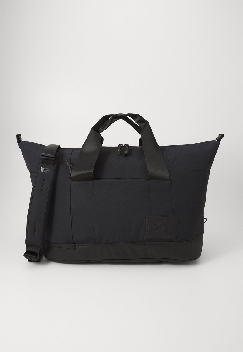 Black duffel bag with nylon material, rectangular shape, dual top handles, adjustable shoulder strap, and logo patch on the side.