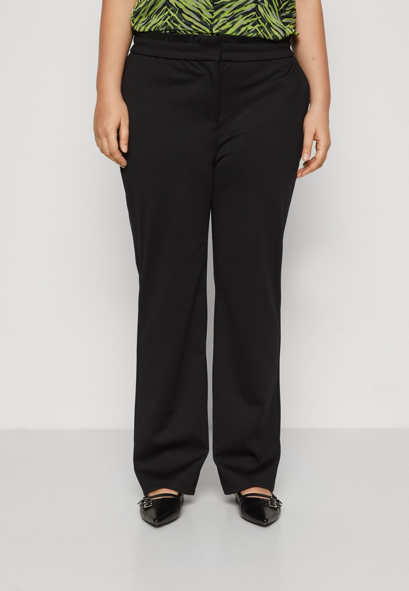 Black, wide-leg trousers with an elastic waistband, paired with a patterned green top. Footwear features pointed toes with decorative buckles.