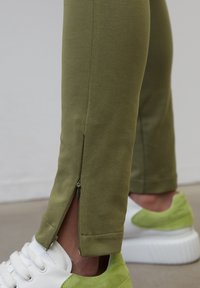 Olivgröna leggings med en slät textur, med en siddragkedja vid vristen. Matchas med vita sneakers med gröna detaljer.