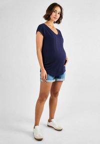 Top premaman a scollo a v con un bel blu navy, in tessuto morbido e dalla vestibilità comoda, abbinato a pantaloni corti in denim chiaro e sneakers bianche con dettagli marroni.