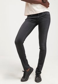 Jeans skinny negros de denim, con un diseño ajustado, cintura a media altura y sutiles detalles de costura. Combinados con zapatos negros de cordones.