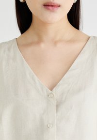 Linen bluza z V-izrezom v svetlo bež barvi, z gumbom za zapiranje in subtilno teksturo. Gladek material s preprostim, minimalističnim dizajnom.