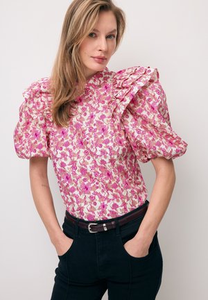 Kobieta o długich blond włosach, ubrana w różową, kwiecistą bluzkę z bufkowymi rękawami oraz ciemne spodnie, z rękami w kieszeniach, stojąca na jednolitym tle.