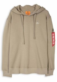 Haut à capuche beige en matériau doux, avec une grande capuche, des cordons de serrage à l'avant, des poignets et un ourlet côtelés, et une poche zippée sur le côté avec une étiquette rouge.
