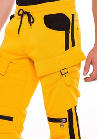 Gelbe Cargo-Hosen mit schwarzen Reißverschlüssen und Seitenschlägen. Zu den Merkmalen gehören Seitentaschen, ein Schnallenriemen und ein Kordelzugbund.