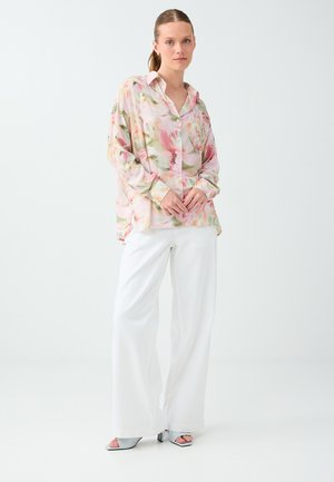 Femme debout portant un chemisier ample à fleurs rose pastel, un pantalon large blanc et des sandales à talons ouvertes sur un fond clair uni.