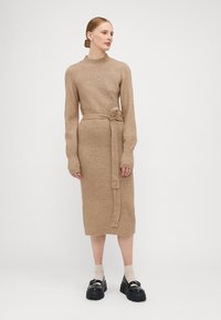 Abito midi lavorato a maglia beige, con collo alto, maniche lunghe e una cintura in vita. Tessuto testurizzato e silhouette dritta, abbinato a scarpe nere chunky.