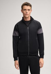 Zwarte zip-up jas met een hoge kraag, voorzien van grijze geometrische schouderaccenten en een getailleerd ontwerp. Gemaakt van gladde stof.