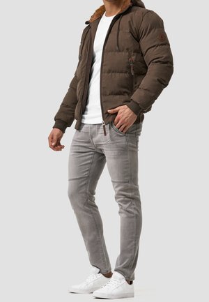 Mann trägt braune gesteppte Jacke mit pelzgefütterter Kapuze, weißes Hemd, hellgraue Jeans und weiße Turnschuhe, steht mit der Hand in der Tasche.