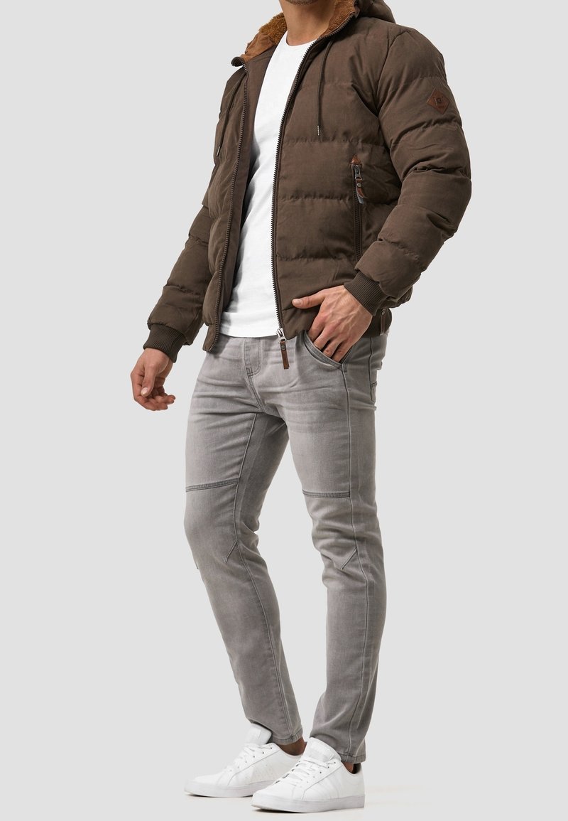 Mężczyzna w brązowej pikowanej kurtce z futrzanym kapturem, białej koszuli, jasnoszarych dżinsach i białych tenisówkach, stojący z ręką w kieszeni.