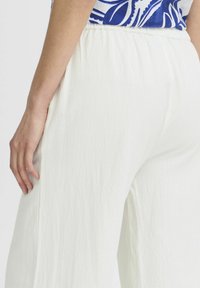 Pantalons larges blancs texturés avec une taille élastique. Couture latérale visible et tissu doux et léger. Surface lisse avec de subtiles irrégularités.