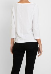 Frau in einem weißen Langarmshirt und schwarzen engen Hosen, von hinten vor einem schlichten hellen Hintergrund betrachtet.