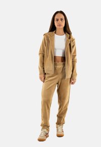 Chándal de pana color marrón con hoodie de cremallera y pantalones con cintura elástica. Llevado con un top corto blanco y zapatillas marrones con acentos rojos.