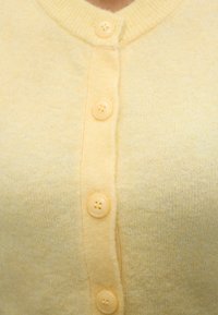 Κοντινό ενός απαλού κίτρινου ζακέτας με τέσσερα ταιριαστά στρογγυλά κουμπιά κλεισμένα μπροστά.