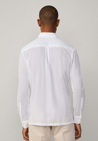 Biała koszula z długim rękawem i kołnierzykiem zapinanym na guziki, wykonana z lekkiego materiału, z plisą z tyłu i mankietami, noszona na jasnych spodniach.