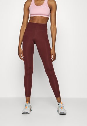 Burgundy high-waisted leggings with a smooth texture and Nike logo; paired with a light pink sports bra and grey athletic shoes with orange accents.