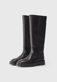 Magas fekete bőr csizmák vastag talppal és finom varrással, egy sima világos háttérrel szemben állva.