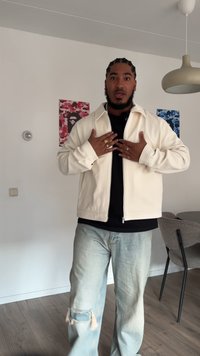 Fényes színű, cipzáros dzseki, textúrált anyagból, fekete belső réteggel, elnyűtt világoskék farmernadrággal párosítva. Nincsenek látható logók vagy minták.