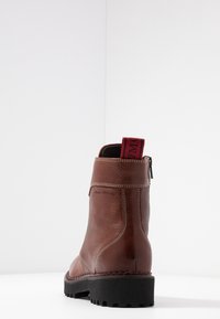 Stivale alla caviglia in pelle marrone con suola nera a cingolo, cuciture bianche, zip posteriore e nastro rosso con testo nero su sfondo bianco.