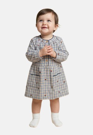 Tout-petit debout, souriant, portant une robe à manches longues à carreaux avec de petites impressions florales et des chaussettes blanches, sur un fond uni.