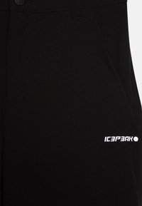 Pantalons en tissu extensible noir avec une texture lisse, poches latérales et un imprimé logo blanc sur le bas à gauche.