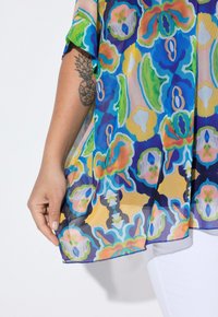 Top trasparente con un vivace motivo floreale multicolore in blu, verde, giallo e arancione. Include maniche ampie e un orlo arrotondato.