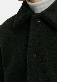 Zelená fleecová bunda s texturovaným povrchem, s špičatým límcem a hnědými knoflíky, která vyzařuje měkký a teplý design.