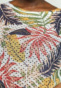 Top ligero de punto abierto con un patrón floral en rojo, verde, amarillo y azul marino sobre un fondo blanco. Presenta un escote amplio.