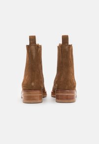 Un par de botas de ante marrón hasta el tobillo con tacones de madera apilada, mostradas desde atrás sobre un fondo blanco.