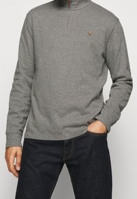 Grå kvartslång zip-tröja i bomullsmaterial, med en liten logotyp på bröstet och ribbade ärmavslut, tillsammans med mörka jeans.